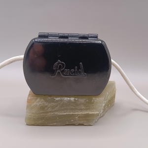Pode incluir: Uma caixa cinza escuro, articulada, com a palavra "Racid" em escrita cursiva. A caixa está apoiada em uma pedra verde clara e texturizada. Um cordão branco está preso à caixa.