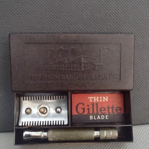 Könnte beinhalten: Ein braunes Plastiketui mit einem Rasierer und einer Packung Gillette Thin Klingen. Das Etui zeigt ein erhabenes Bild einer Fabrik und den Text "Mofem Onkordtvakeszulek".