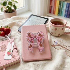 Op de afbeelding: Een roze tablethoes met een doorzichtige strikvormige telefoonhouder met bruine teddyberen en aardbeien. Een mok met aardbeienpatroon, een kom met aardbeien en een roze notitieboek zijn ook zichtbaar.