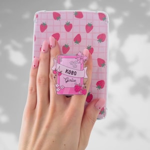 Könnte beinhalten: Eine Hand mit rosa Nägeln hält eine pinkfarbene Handyhülle mit Erdbeermuster. Ein pinkfarbener, buchförmiger Handyhalter mit der Aufschrift "KOBO Girlie" ist befestigt. Der Halter zeigt Erdbeeren, Blumen und eine Schleife.