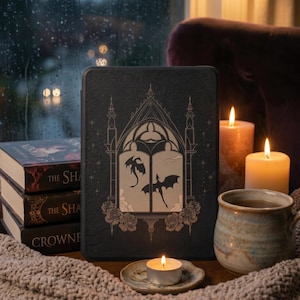 Op de afbeelding: Een zwarte e-reader hoes met een goudkleurige illustratie van een gotisch boogvenster met twee vliegende draken en rozenaccenten. De hoes staat naast een stapel van drie boeken, een keramische mok en twee brandende kaarsen op een ondergrond.