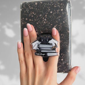Può includere: Un supporto per telefono in acrilico bianco e nero con un design a forma di drago dei cartoni animati, ali bianche e una pila di libri. I libri hanno le parole "book dragon" stampate sopra. La custodia del telefono dietro il supporto ha uno sfondo nero con un motivo floreale.
