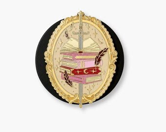 May include: A circular enamel pin featuring a gold ornate oval frame surrounding a stack of books pierced by a silver sword. The books are colored in shades of pink and red, with a crescent moon symbol on the red spine, accented by floating feathers.