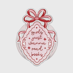May include: A pink and red decorative badge with the text "girls just wanna read books." The badge has a scalloped edge and a red bow at the top. The text is in a handwritten style.