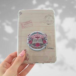 Peut inclure: Coque de téléphone transparente avec un motif de lettre vintage et un autocollant "Romantasy Reader Society". L'autocollant représente un livre ouvert, des roses et un dragon. La coque est tenue par une main avec une manucure rose.