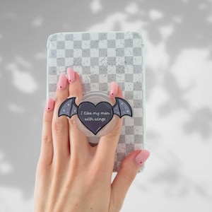 Op de afbeelding: Een doorzichtige telefoonhouder met een hartvormig ontwerp en vleermuisvleugels. De tekst "I like my men with wings" staat op het hart. De telefoonhoes heeft een grijs-wit geblokt patroon.