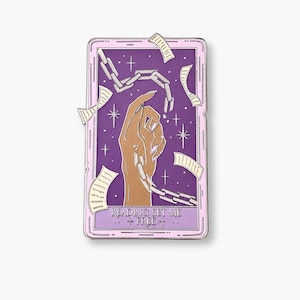 May include: A rectangular enamel pin featuring a purple background with a tan hand breaking free from silver chains. Floating paper scraps surround the hand, and the text READING SET ME FREE is printed at the bottom. The pin has a light pink border.