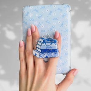 Può includere: Un supporto per telefono in acrilico trasparente con una pila di libri e le parole "Bookish Girlie" in blu. Il supporto ha un design a fiocco ed è posto contro una custodia per telefono azzurra con un motivo floreale e a fiocco.