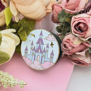 Può includere: Un supporto rotondo per telefono pop-up azzurro con un design a castello stravagante con torrette viola e dettagli dorati. Il design include nuvole, stelle e creature volanti. L'impugnatura ha un bordo dorato e una base verde scuro.