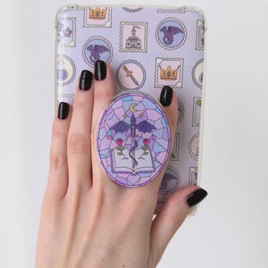 Puede incluir: Una funda transparente para teléfono con un diseño de sellos morados con dragones, coronas y castillos. Un soporte redondo y morado para teléfono desplegable con un diseño de vidriera de un dragón, espada, rosas y un libro abierto está adherido a la funda.