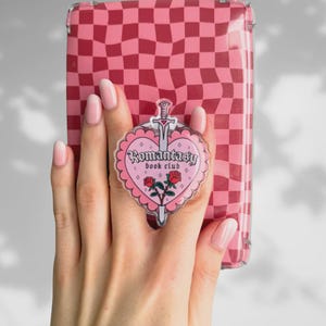 Può includere: Una custodia per telefono a scacchi rosa e rossi con un supporto per telefono a forma di cuore. L'impugnatura presenta il testo "Romantasy book club" con un design a spada e rose. La custodia del telefono è tenuta da una mano con smalto rosa.