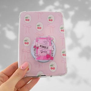 May include: A pink Kindle case with a repeating pattern of cherries and bows. A circular sticker with the words "Kindle Girlie" and illustrations of flowers, strawberries, and a pink Kindle is attached to the case.