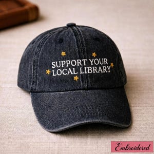 Peut inclure: Casquette de baseball gris foncé avec le texte brodé "SUPPORT YOUR LOCAL LIBRARY" en blanc, orné d'étoiles dorées. La casquette a une visière incurvée et un style classique et décontracté.