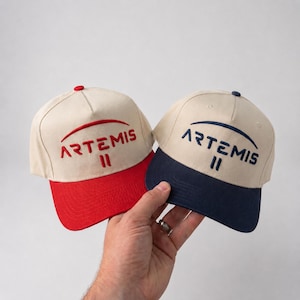 Könnte beinhalten: Zwei beige Baseballkappen mit dem Wort "ARTEMIS" auf der Vorderseite gestickt. Eine Kappe hat einen roten Schirm und rote Stickerei, die andere einen marineblauen Schirm und Stickerei. Die Kappen werden in einer Hand gehalten.