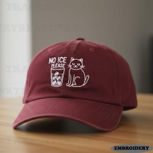 Puede incluir: Gorra de béisbol granate con visera curva. La gorra presenta texto bordado en blanco que dice "NO ICE PLEASE" junto a un gato de dibujos animados y un vaso de cubitos de hielo. La gorra está hecha de tela suave.