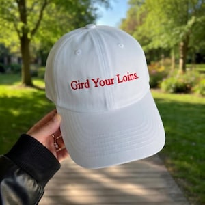 Puede incluir: Gorra de béisbol blanca con la frase bordada en rojo "Gird Your Loins". La gorra tiene una visera curva y una corona estructurada. El fondo muestra un entorno de parque verde borroso.