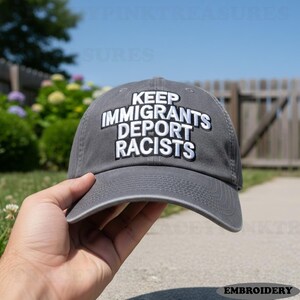 Puede incluir: Gorra de béisbol gris con la inscripción bordada en blanco "KEEP IMMIGRANTS DEPORT RACISTS". La gorra tiene una visera curva y está hecha de una tela suave. El fondo muestra una valla y vegetación.
