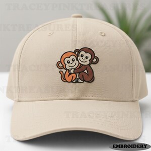 Può includere: Cappellino da baseball beige con un disegno ricamato di due scimmie dei cartoni animati che si abbracciano. Una scimmia è arancione, l'altra marrone. Il cappellino ha una visiera curva e un bottone sulla parte superiore. La parola "EMBROIDERY" è stampata in basso a destra.