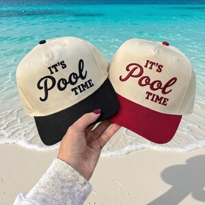 Op de afbeelding: Twee baseballpetten met de tekst "IT'S Pool TIME" geborduurd op de voorkant. Eén pet heeft een zwarte klep, de andere een bordeauxrode klep. De petten zijn beige.
