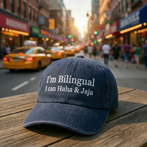May include: A navy blue baseball cap with white embroidered text reading "I'm Bilingual I can Haha & Jaja". The cap rests on a wooden surface, with a city street and yellow taxis visible in the background.