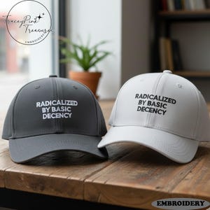 Op de afbeelding: Twee baseballpetten, één donkergrijze en één witte, worden getoond op een houten oppervlak. Beide petten hebben de tekst "RADICALIZED BY BASIC DECENCY" in het wit geborduurd. De petten maken deel uit van een collectie geborduurde hoeden.