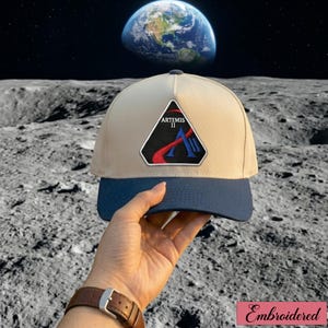 Op de afbeelding: Beige en marineblauwe baseballcap met het Artemis II-logo. Het logo is een zwarte driehoek met rode en blauwe accenten en de tekst "ARTEMIS II". De cap heeft een gebogen klep en een donkerblauwe klep.
