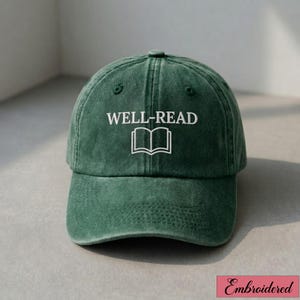 May include: A dark green baseball cap featuring the words "WELL-READ" and an open book graphic embroidered in white. The cap has a curved peak and a classic six-panel design, suitable for casual wear.