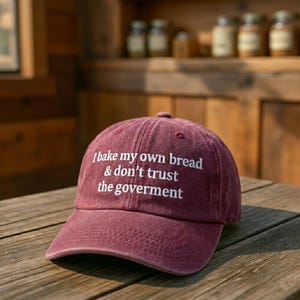 Può includere: Cappellino da baseball bordeaux con testo bianco ricamato: "I bake my own bread & don't trust the government". Il cappellino è su una superficie di legno. Il cappello ha una visiera curva e un design a sei pannelli.