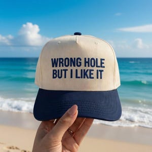 May include: A cream-colored baseball cap with a navy blue brim. The cap features the embroidered text "WRONG HOLE BUT I LIKE IT" in navy blue. The cap is held up against a backdrop of a beach and ocean.