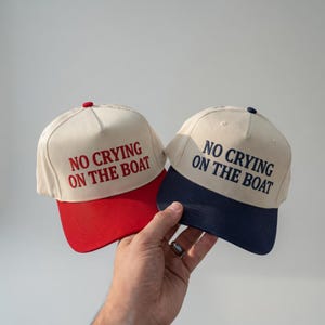 Puede incluir: Dos gorras de béisbol con el texto "NO CRYING ON THE BOAT". Una gorra tiene una visera roja y la otra una visera azul marino. Ambas gorras tienen una corona color crema y un botón en la parte superior.