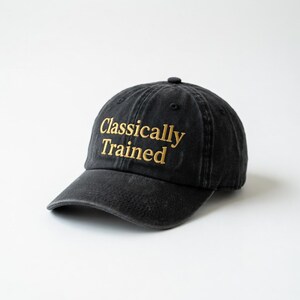 May include: Black baseball cap with a curved brim. The cap features the embroidered text "Classically Trained" in gold thread. The hat is made of a soft, worn material and has a classic six-panel design.