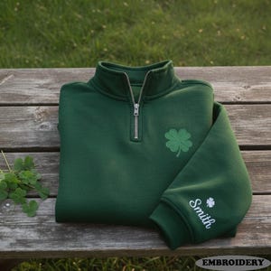Könnte beinhalten: Ein gefalteter dunkelgrüner Sweatshirt mit einem Viertel-Reißverschlusskragen und einem Kleeblatt auf der Brust. Der Ärmel zeigt den Namen "Smith" und ein kleines Kleeblatt. Das Sweatshirt liegt auf einer Holzoberfläche.