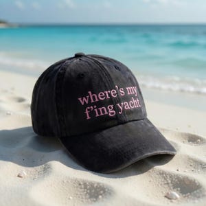 May include: A black baseball cap with the embroidered text "where's my f'ing yacht" in pink. The cap is sitting on a sandy beach with the ocean in the background.