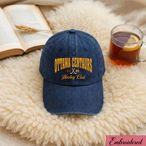 May include: A blue denim baseball cap with the text "OTTAWA CENTAURS Hockey Club" in gold lettering. The cap has a curved brim and a classic six-panel design. The text includes the numbers "24" and "81" with crossed hockey sticks.