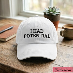 Pode incluir: Boné de beisebol branco com o texto bordado "I HAD POTENTIAL" em preto. O boné está sobre uma superfície de madeira. Uma pequena planta suculenta num vaso de terracota e uma chávena de café estão no fundo.
