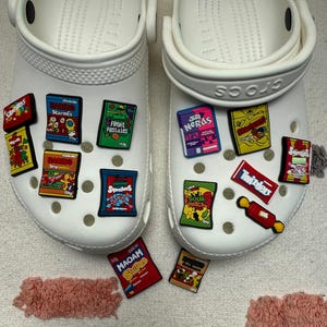 May include: A pair of white clogs decorated with various colourful candy-themed charms. The charms feature logos and packaging designs for brands including Haribo Starmix, Tangfastics, Fruit Pastilles, Squashies, Nerds, Skittles, Twizzlers, and Maoam.