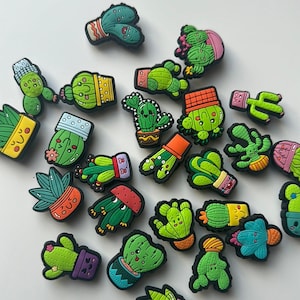 May include: A collection of small, colourful rubber shoe charms shaped like various cacti in pots. Each charm features a unique green cactus design with a smiling face, set in a brightly coloured planter. The charms are scattered across a plain surface.