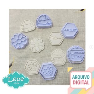 May include: A collection of light blue and off-white cookie cutters in various shapes, including flowers, hexagons with text, and a bee. The image also includes the text "Lepe Scrap" and "ARQUIVO DIGITAL".