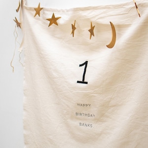 May include: A white fabric banner with gold stars and a crescent moon hanging from the top. The banner says "Happy Birthday Banks" and has the number "1" in the center.
