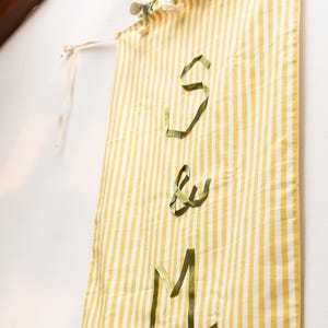 May include: A yellow and white striped banner with the letters "S & M" made of green ribbon. The banner is decorated with white flowers and is hanging on a white wall.