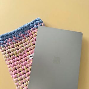 Könnte beinhalten: Ein grauer Laptop liegt auf einer gehäkelten Laptoptasche in Rosa-, Gelb- und Blautönen. Die Tasche hat ein Etikett mit dem Wort "par". Der Hintergrund ist einfarbig gelb. Der Laptop ist ein modernes Design.
