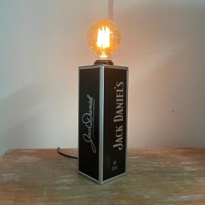 Può includere: Una lampada da tavolo nera e argento con le parole "Jack Daniel's" stampate sul davanti. La lampada ha una base rettangolare e una lampadina rotonda e luminosa sulla parte superiore. La lampada è collegata a una presa elettrica.