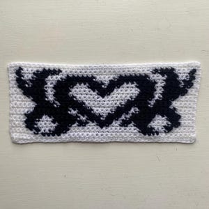 May include: A rectangular, handmade crochet piece featuring a black heart and XOXO design on a white background. The stitches are clearly visible, showcasing the texture of the yarn. The piece is likely a decorative item or part of a larger craft project.