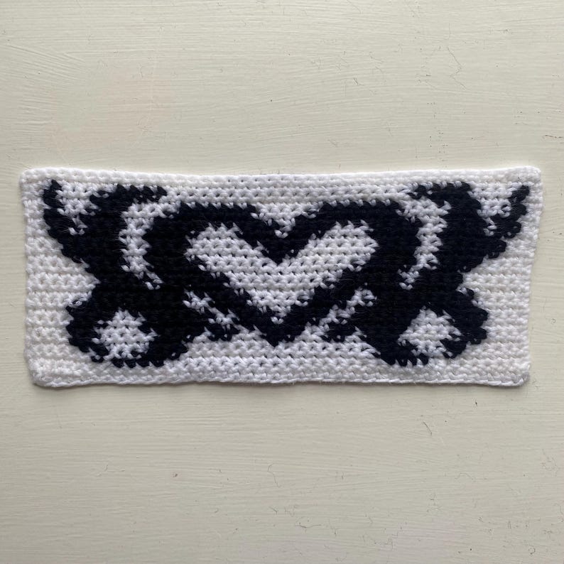 May include: A rectangular, handmade crochet piece featuring a black heart and XOXO design on a white background. The stitches are clearly visible, showcasing the texture of the yarn. The piece is likely a decorative item or part of a larger craft project.