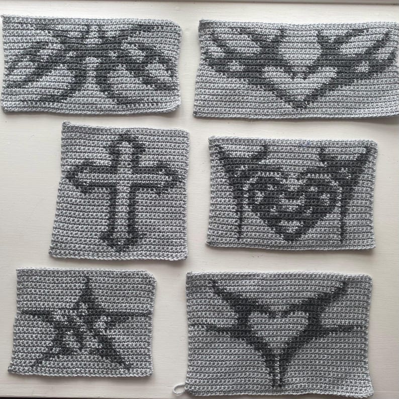 May include: Six gray crocheted squares with different black designs. The designs include a cross, a heart, a butterfly, and a star.