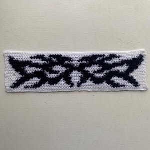 May include: A white crocheted headband with a black geometric design. The headband is rectangular and features a repeating pattern of zigzags and angular shapes. The design is centered on the headband.