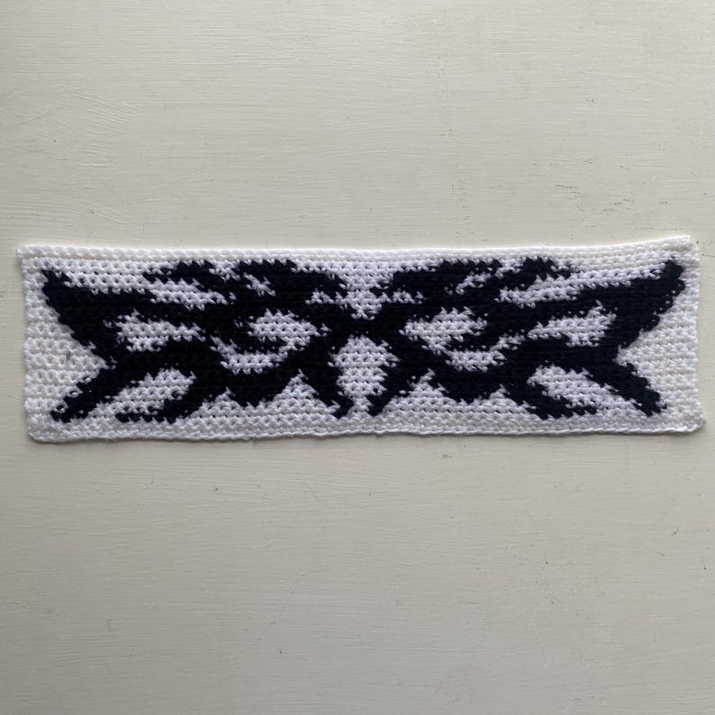 May include: A white crocheted headband with a black geometric design. The headband is rectangular and features a repeating pattern of zigzags and angular shapes. The design is centered on the headband.