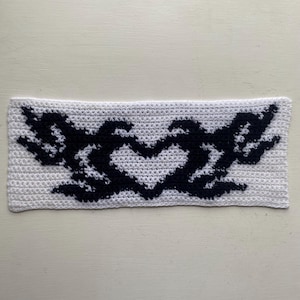 May include: A white crocheted headband featuring a black heart-shaped design with wing-like elements on either side. The handmade accessory is crafted with a textured stitch, showcasing a contrasting color pattern.