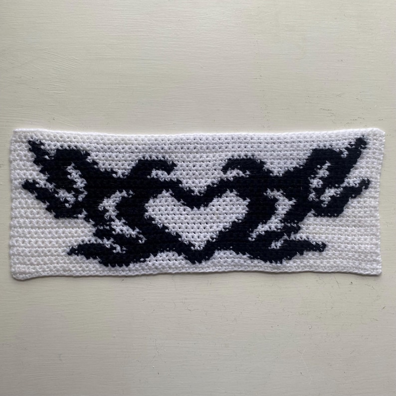 May include: A white crocheted headband featuring a black heart-shaped design with wing-like elements on either side. The handmade accessory is crafted with a textured stitch, showcasing a contrasting color pattern.