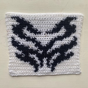 May include: A square, white crocheted item with a black, wing-like design in the center. The design is symmetrical and detailed, with the black sections resembling feathers or stylized wings. The item is handmade and has a textured surface.
